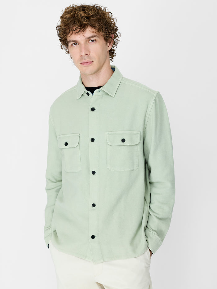 A man wearing a sage green over shirt with long sleeves. a pointed collar and a button down fastening.