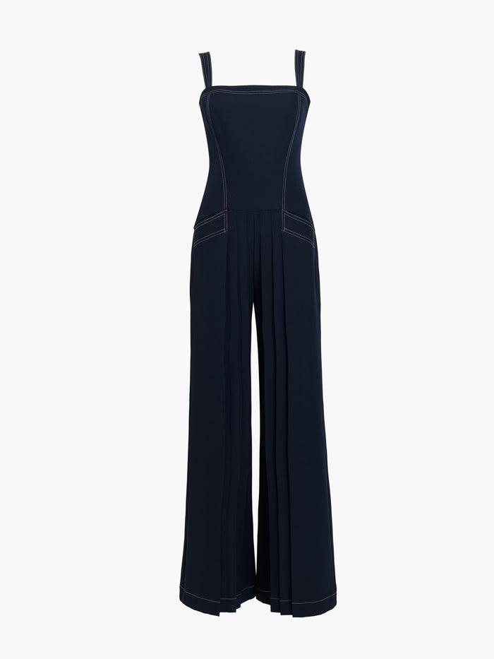 Woman wearing a pleated navy blue full length wide leg jumpsuit with straps, a square neckline, front pockets, dropped waist and contrast stitching.