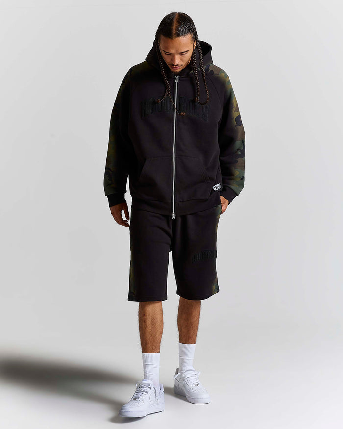 A man modeling a black zip-up hoodie with dark green camouflage sleeves and hood, matching black shorts, white socks, and white sneakers, shot against a light gray studio background.
