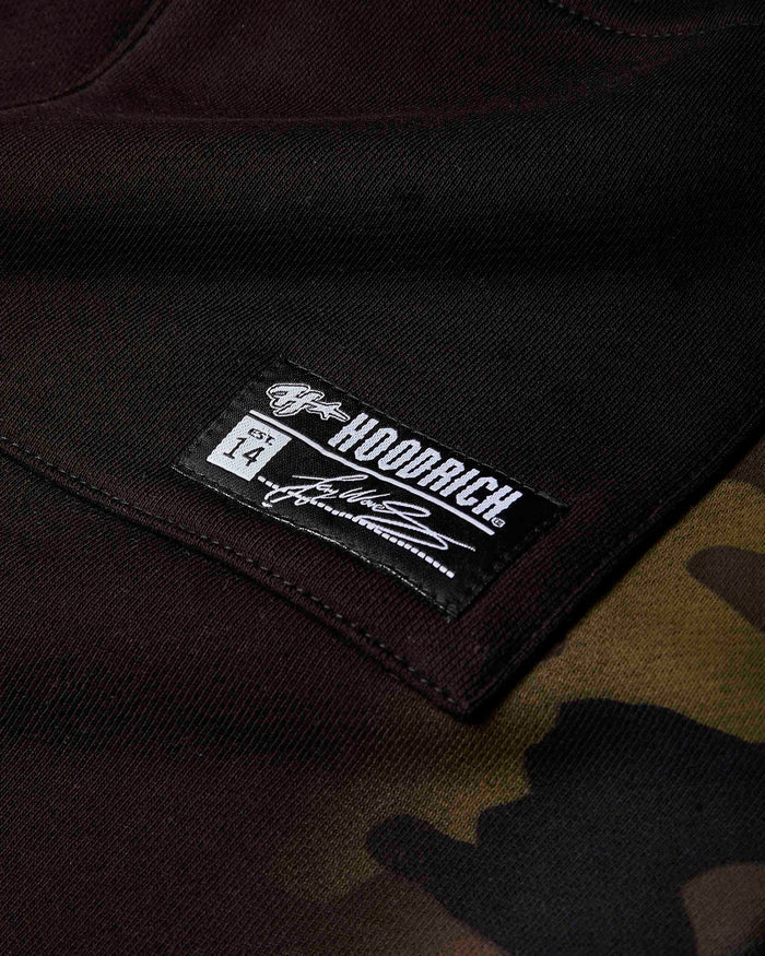 Close-up of a black HOODRICH brand patch sewn onto a dark brown and black camouflage hoodie.