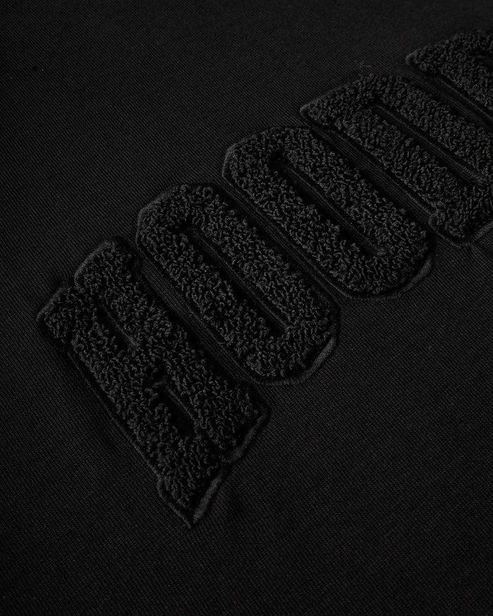 Close-up of a black textured fabric with large raised embroidered letters reading 'HOODRICH' in black-on-black thread.