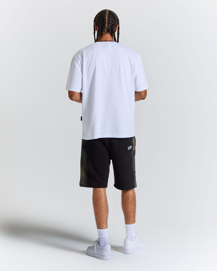 A man viewed from behind wearing a white t-shirt, black shorts with green camouflage side panels, white socks, and white sneakers, standing against a light gray studio background.