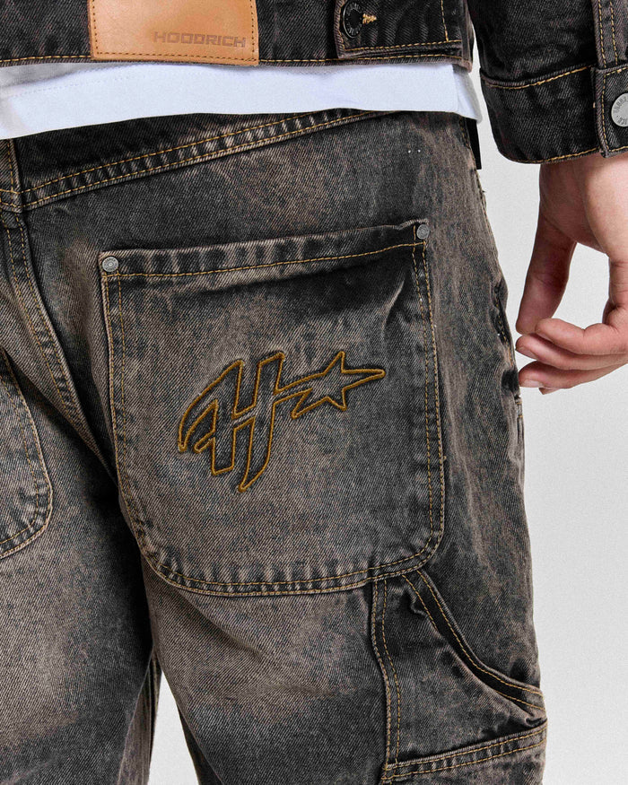 Close-up of the Hoodrich logo embroidered on the pocket of the regular fit black rust washed denim jorts worn by a model.