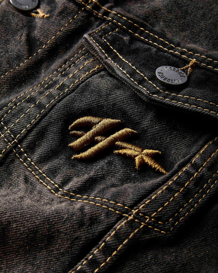 Close-up of the Hoodrich logo embroidered on the chest pocket of the black rust washed denim jacket.