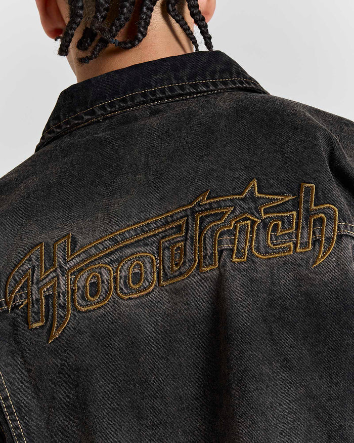 Close-up of the Hoodrich logo embroidered on the chest pocket of the black rust washed denim jacket worn by a model.