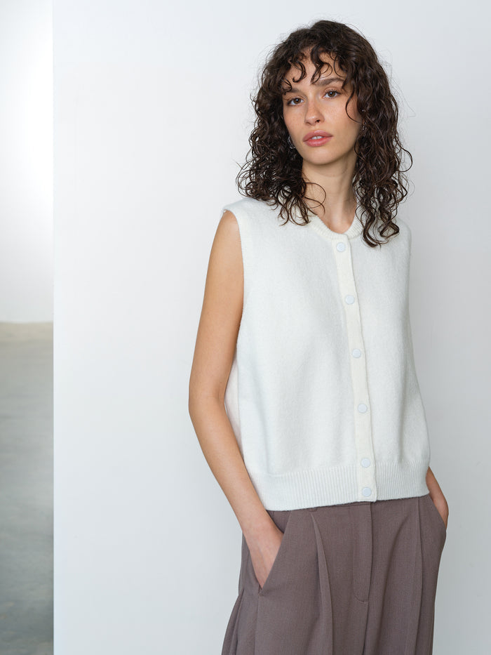 Woman wearing a cream, sleeveless knitted tank top with a round neckline and button down fastening