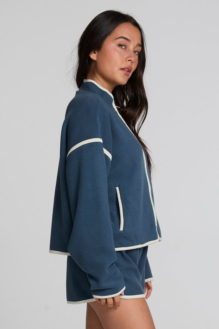 Side view of a woman wearing the Chaser Sharun Dark Denim Jacket and shorts on a neutral background