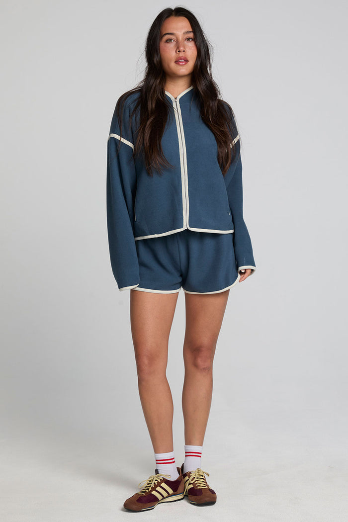 Full body front view of a woman wearing the Chaser Sharun Dark Denim Jacket and matching blue shorts on a neutral background