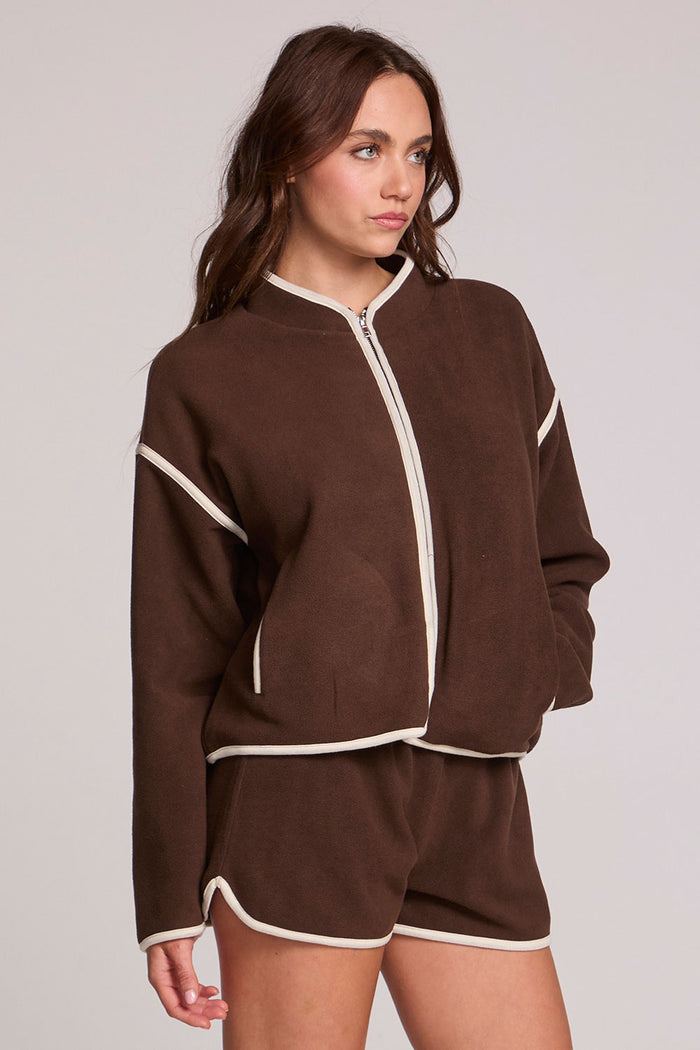 Side angle view of a woman wearing the Chaser Sharun Worn Brown Jacket on a neutral background with matching shorts