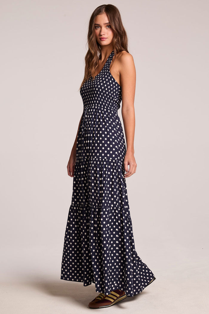 Side angle view of a woman wearing the Chaser  Siren Navy Spot Maxi Dress on a neutral background with one foot raised