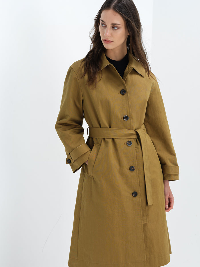 A woman wearing a khaki longline belted trench coat with long sleeves, a pointed collar and button down fastening.