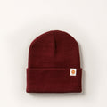 The Stretch Cuff Beanie- Burgundy Red is a maroon acrylic beanie with a stretch cuff and a small white patch featuring a brown animal graphic on the front, shown against an off-white background.