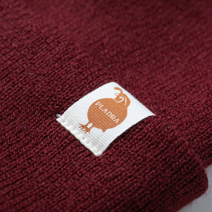 Close-up of the Stretch Cuff Beanie in Burgundy Red, featuring a white rectangular label with an orange bird and the word PEADRA.
