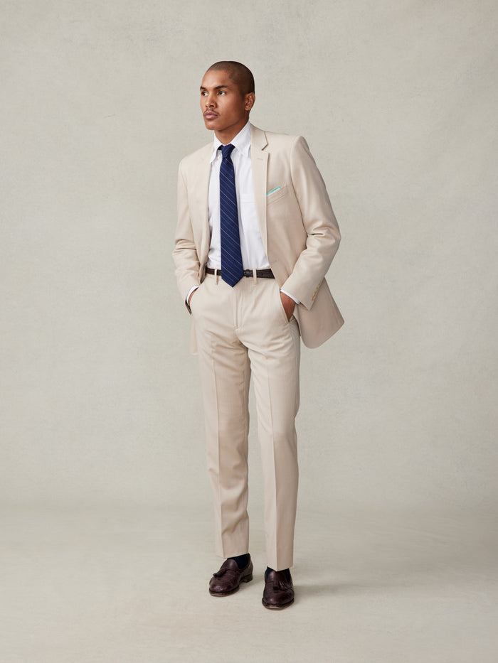 Man in tan suit with hands in pockets, white shirt, navy tie, front full-body view