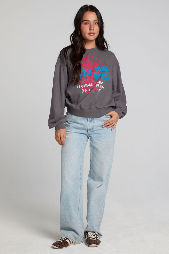 Full body view of a woman wearing the Chaser The Paris Punk Graphic Sweatshirt and jeans on a neutral background with arms lowered and one hand on hip