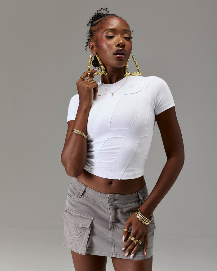 Woman wearing a white top and gray skirt with gold jewelry against a gray background