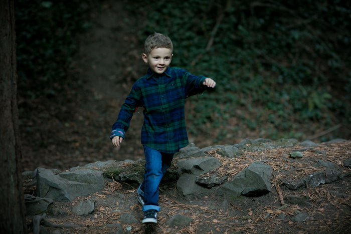 A young boy in a Pladra Toddler Cubs Flannel Shirt - Bolinas Blue smiles while walking on rocky ground in a forest.