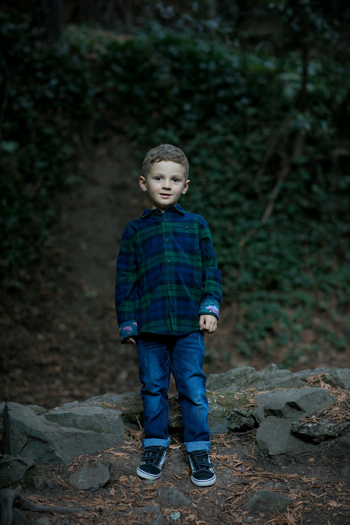 Young boy wears Pladra Toddler Cubs Flannel Shirt in Bolinas Blue, standing on rocks in a wooded, green area.