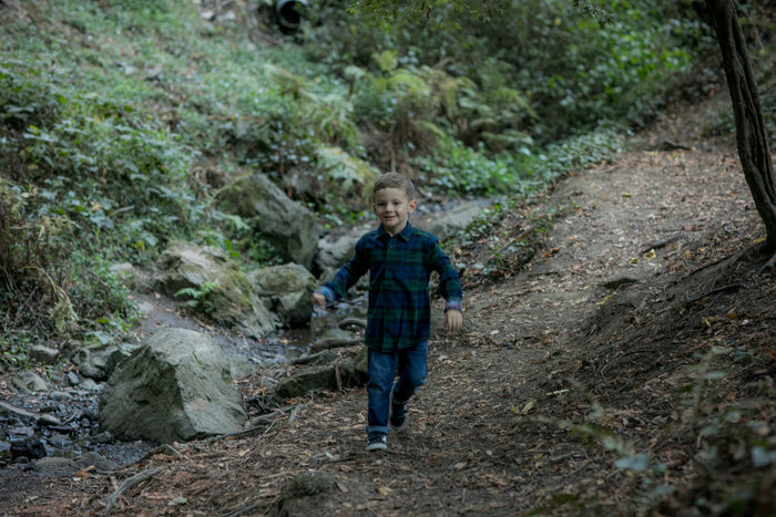 A young boy in a Pladra Toddler Cubs Flannel Shirt—Bolinas Blue smiles as he walks a forest trail amid rocks and greenery.
