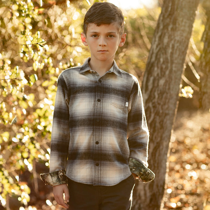 A boy wears the Pladra Toddler Cubs Flannel Shirt in Sterling Grey Heather outdoors among trees and sunlight.