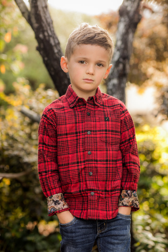 Young boy in a Pladra Toddler Cubs Flannel Shirt—Wrangler Red—stands outdoors, hands in pockets, looking serious.