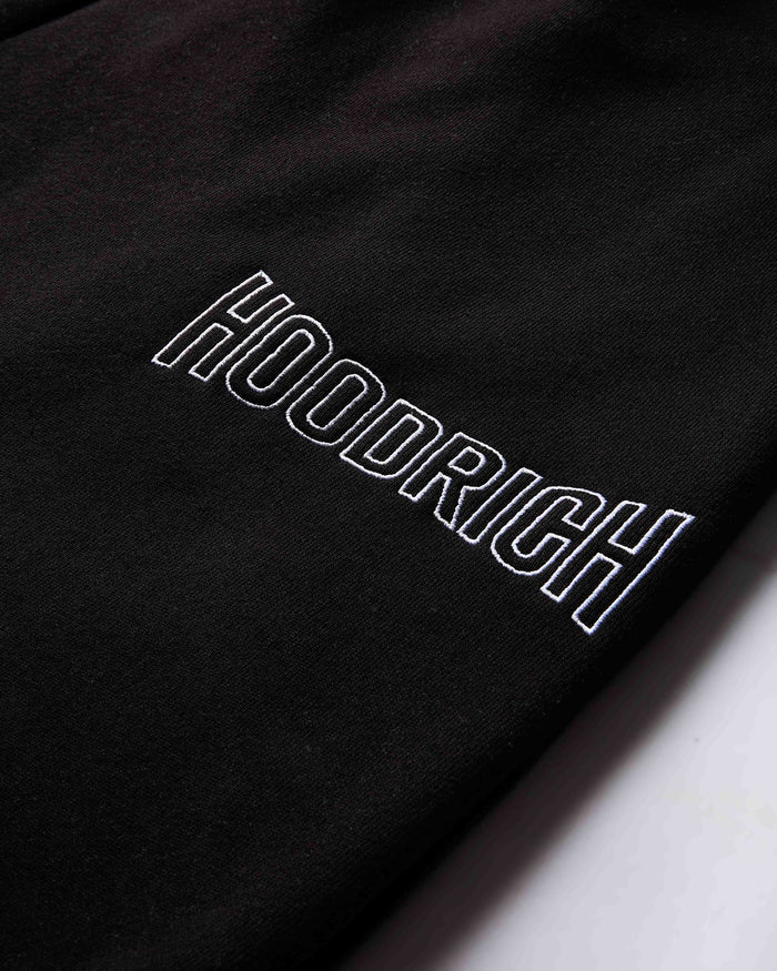 Close-up of the white outlined Hoodrich logo embroidered on the black fabric of the joggers.