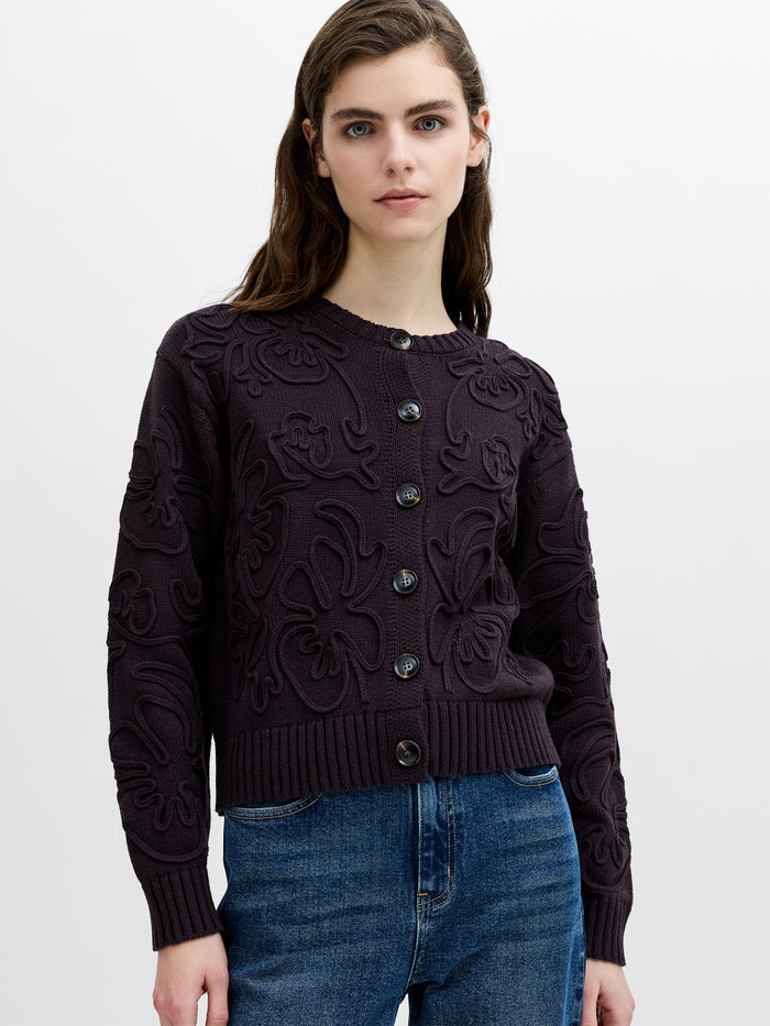 A woman wearing a dark brown 3D box fit, floral knit cardigan with button through fastening, a ribbed crew neck and long sleeves.