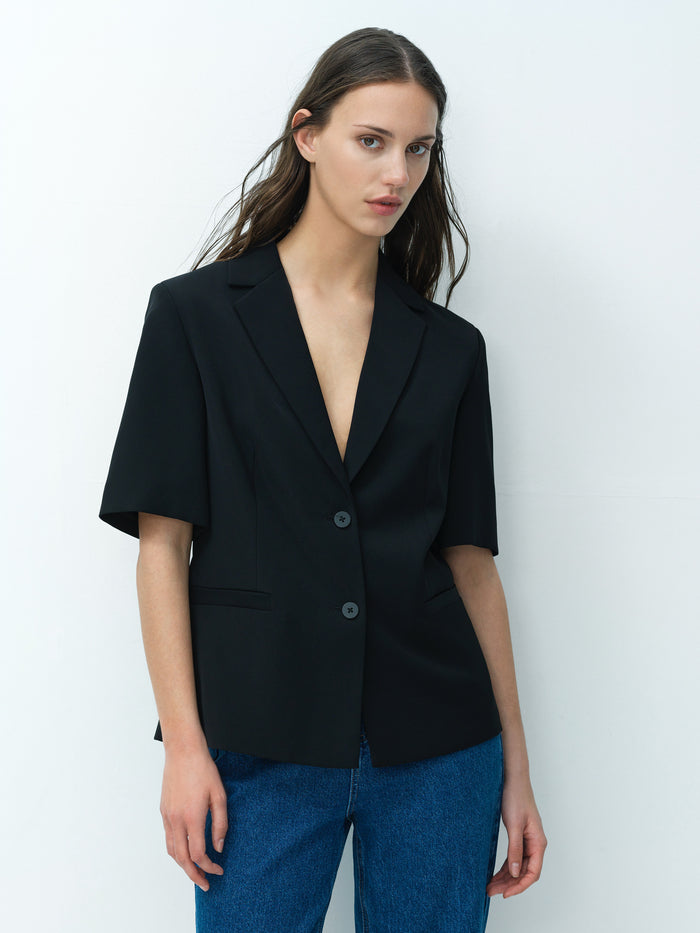 A woman wearing a tailored black short sleeved blazer with a collared V-neckline and 2 front buttons.