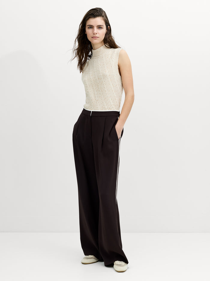 A woman wearing tailored brown wide leg trousers with a cream side trim, front pleats, an elasticated waistband  and a zip and hook fastening.