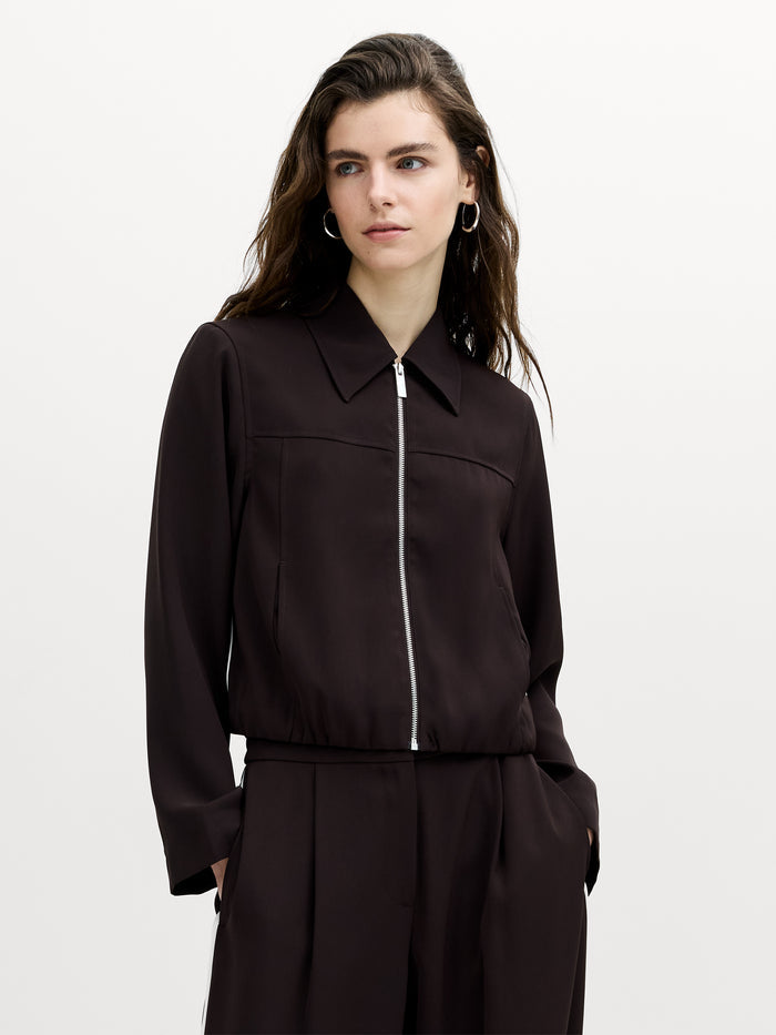 A woman wearing a tailored brown zip front jacket with a pointed collar, contrasting cream side piping and two pockets.