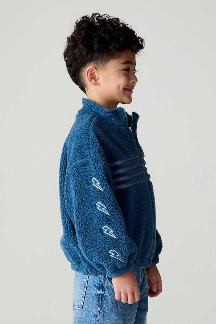 Side view of a boy wearing the Chaser White Bolts Boys Jacket and jeans on a neutral background with arms at sides