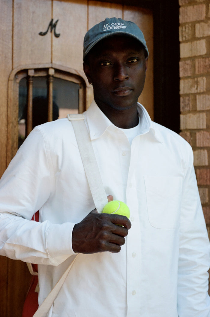 Man in white oxford shirt and cap holding a tennis ball outdoors