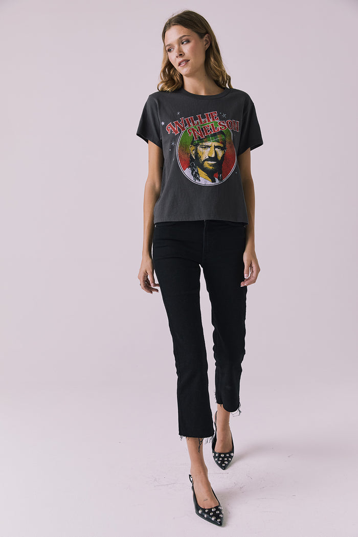 Full body view of woman wearing Chaser’s Willie Nelson Willie Christmas Tee with colorful graphic design and blue jeans on a plain background (licensed band collaboration)