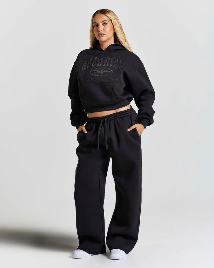A woman modeling a black cropped hoodie and matching wide-leg sweatpants set. The hoodie features a tonal black graphic on the chest with the word 'HOODRICH' and a horse logo.