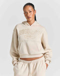A model wearing a cream-colored cropped hoodie with 'HOODRICH' embossed on the chest, paired with matching jogger pants, against a light gray studio background.