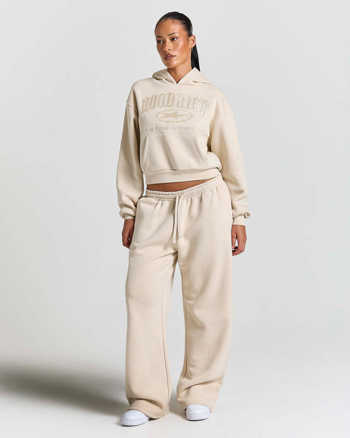 A model wearing a matching beige cropped hoodie and wide-leg sweatpants set with white sneakers, standing against a light gray studio background.