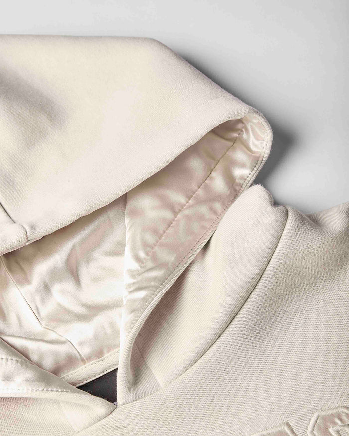 Close-up of a cream-colored hoodie showing the interior satin lining of the hood and collar, featuring a smooth satin-like inner fabric and a textured knit outer fabric.