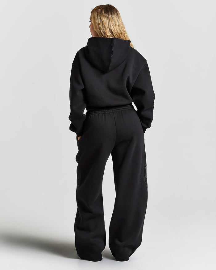 A woman viewed from behind wearing a black cropped hoodie and matching black wide-leg sweatpants against a light gray studio background.