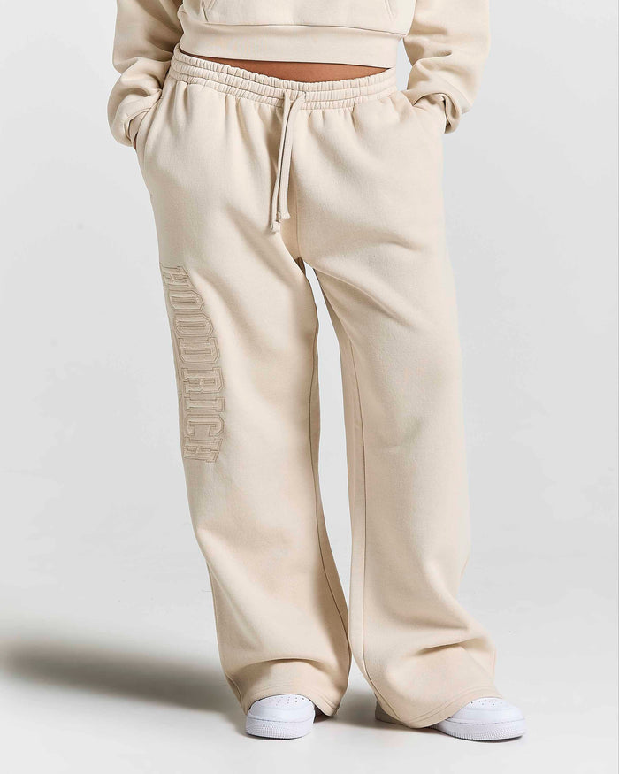 A person wearing cream-colored wide-leg sweatpants with a vertical tonal 'HOODRICH' logo on the right leg, paired with white sneakers against a light gray studio background.