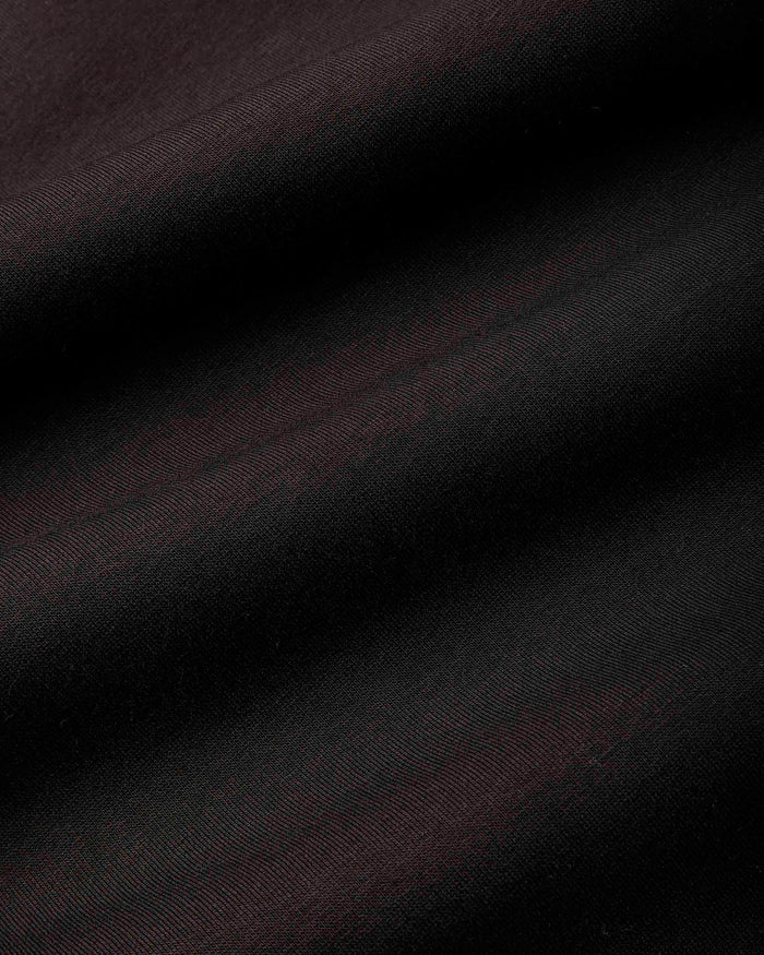 A close-up of dark fabric with a subtle sheen, showing diagonal folds and a fine woven texture in very dark brown or near-black tones.
