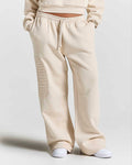 A person wearing cream-colored wide-leg sweatpants with a vertical tonal 'HOODRICH' logo on the left leg, paired with white sneakers against a light gray studio background.