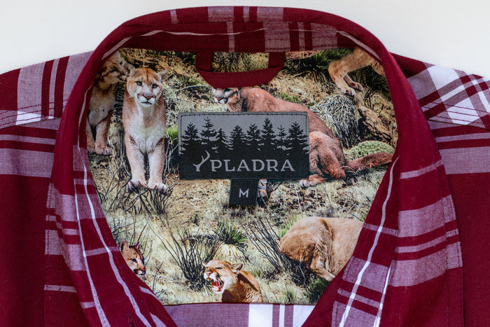 Close-up of a Women's Breeze Shirt- Merlot Red collar in red plaid organic cotton, lined with mountain lion illustrations in grass. The PLADRA label features a forest graphic and size M below.