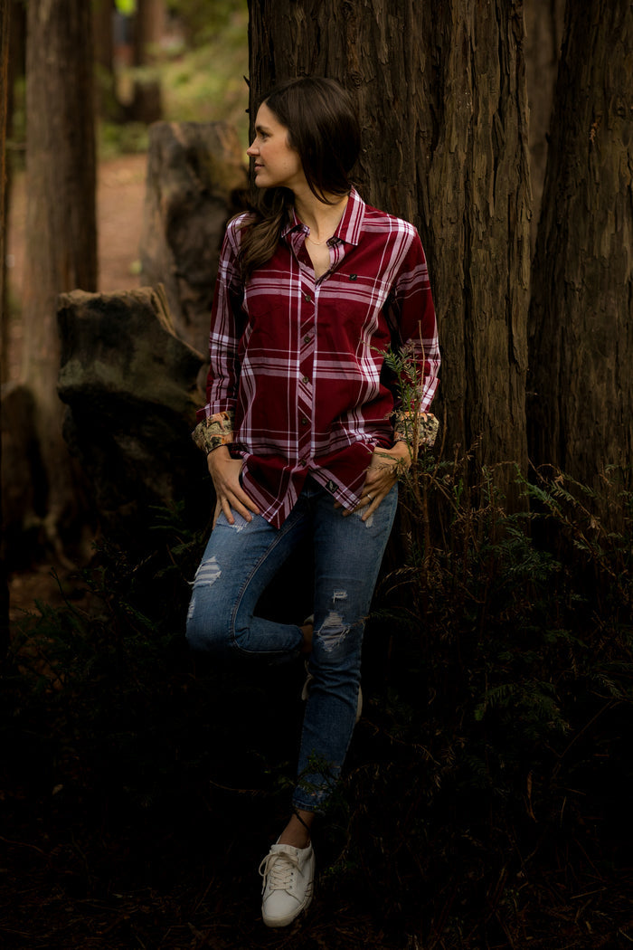 A woman in a Women's Breeze Shirt- Merlot Red, ripped jeans, and white sneakers leans against a tree in a forest, gazing to the side with one leg bent and her hands tucked in her pockets.