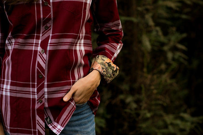 A person wearing the Women's Breeze Shirt – Merlot Red, a lightweight, wrinkle-resistant top with rolled-up sleeves, and blue jeans stands outdoors with greenery blurred in the background.