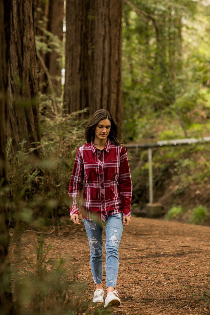 A woman in a Merlot Red Women's Breeze Shirt and ripped jeans walks along a dirt path surrounded by tall trees and lush forest greenery.