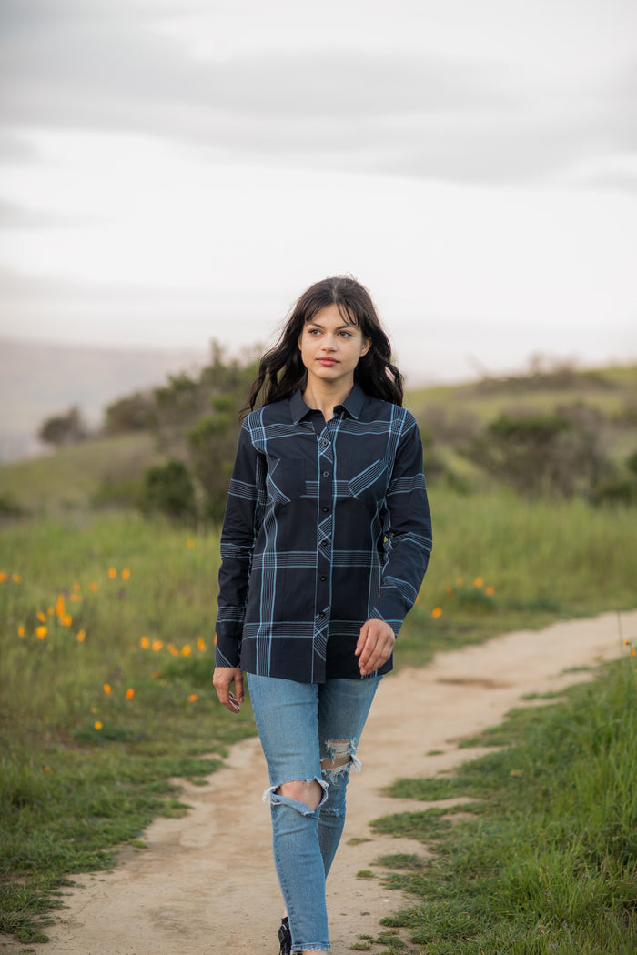 A woman with long dark hair walks along a dirt path in a grassy field with wildflowers, wearing the Women's Breeze Shirt- Ocean Blue and ripped blue jeans. The sky is overcast, with trees visible in the background.