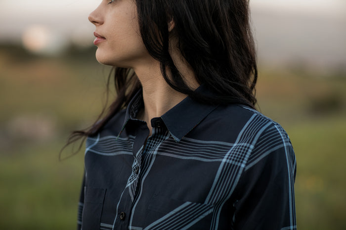 A woman with long dark hair, wearing the Women's Breeze Shirt in Ocean Blue, stands outdoors in a blurred natural setting. The image is cropped at the shoulders with only part of her face visible as she looks to the left.