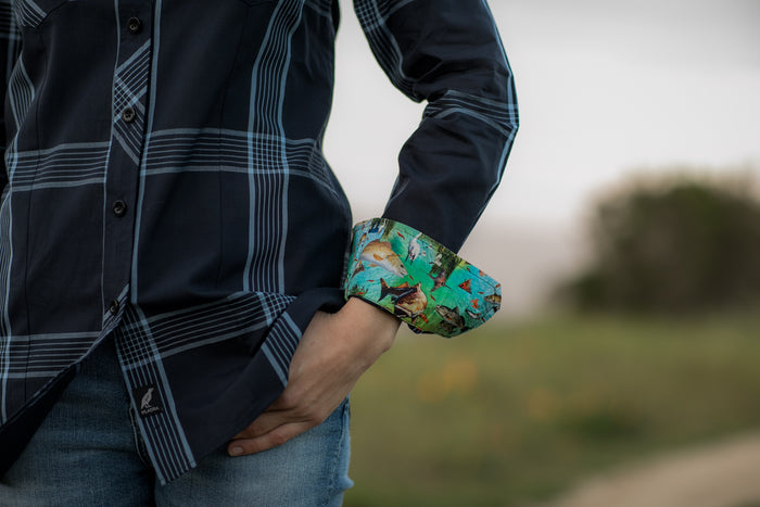 A woman outdoors wears the Women's Breeze Shirt- Ocean Blue with sleeves rolled up, revealing a colorful inner pattern. One hand is in her pocket, paired with blue jeans amid grass and a blurred background.