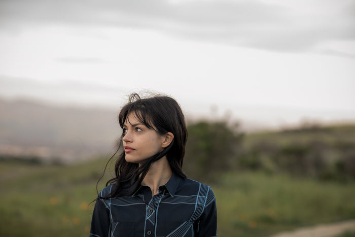 A young woman with dark hair stands in a grassy field under cloudy skies, wearing the Women's Breeze Shirt in Ocean Blue. She looks to her left; distant hills and a blurred background complete the outdoor scene.