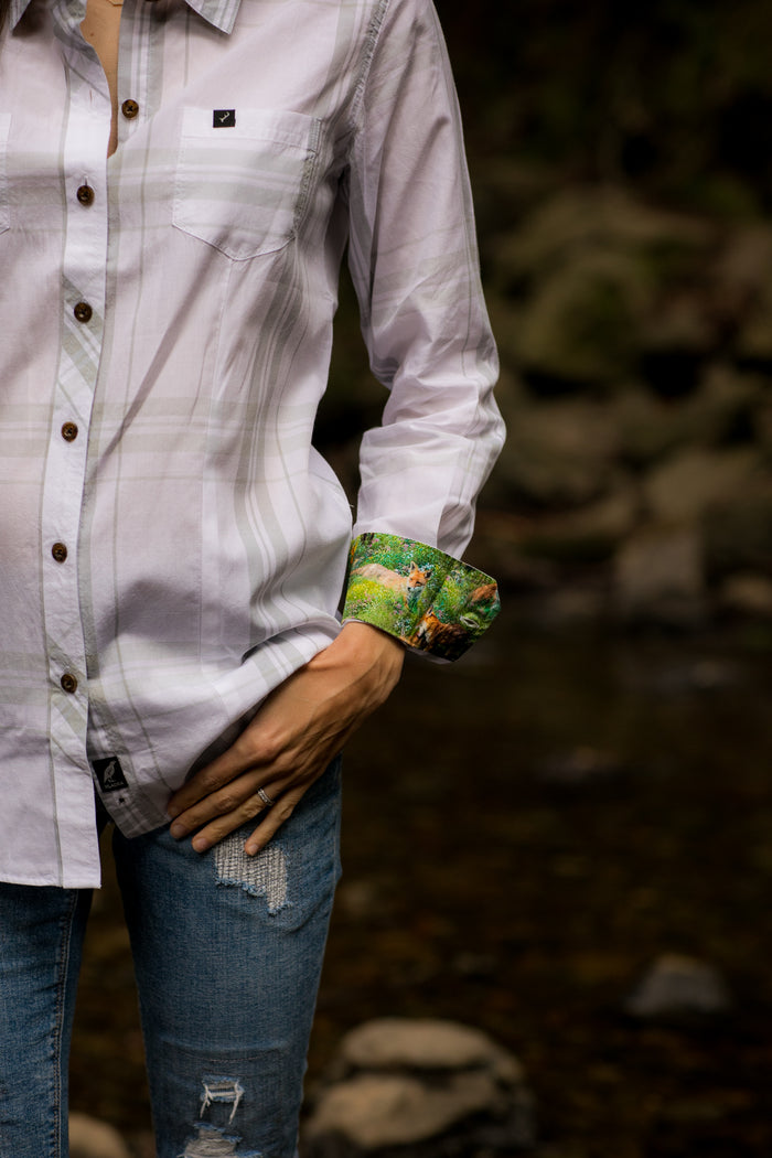 A woman in a Pladra Women's Breeze Shirt–Spring White and blue ripped jeans stands by a stream, hand on hip.